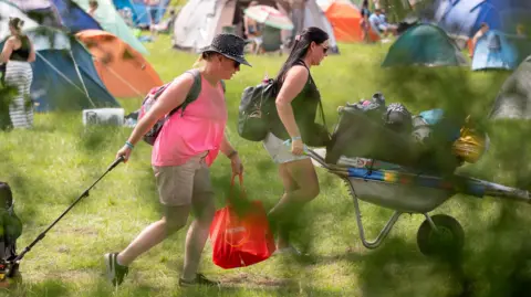 Dylan Roberts Two women wheel camping equipment on a wheelbarrow and a trolley in a field of tents.