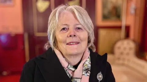 Martin Giles/BBC Penny Carpenter who is sitting down looking and smiling directly at the camera. She has short hair and is wearing a black outdoor coat with a pink scarf.