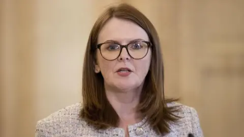 A woman with dark hair is wearing glasses and a grey jacket. She is in mid speech, and behind her is a beige wall. 
