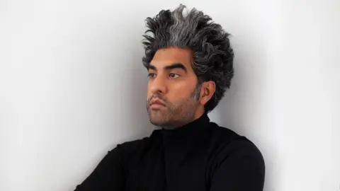 Mahtab Hussain Mahtab has dark and grey hair that is long and wavy, styled to stand upwards. He has dark grey facial hair and is wearing a black top