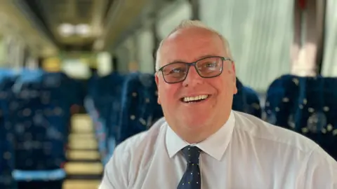 Martin Giles/BBC Charles Sanders is wearing a white shirt and blue tie, which has brown and white dots on it. He is sitting in a passenger seat on a bus. He is also wearing glasses.