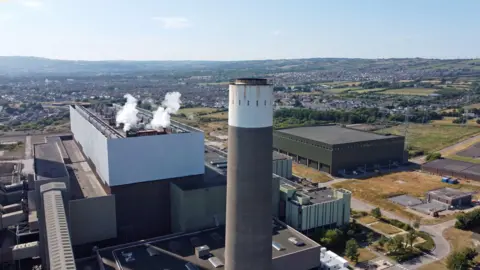 AFP via Getty Images Aerial view of Kilroot - a large grey power station with a chimney. There is smoke leaving the roof of the building and hills with dwellings visible along the horizon.