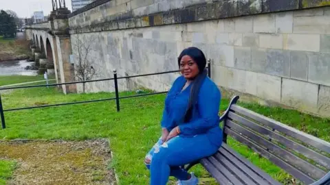 Cynthia Waggah sitting on a wooden bench. She has long black hair and is wearing a blue shirt and blue ripped jeans. The bench is beside a bridge over a river.
