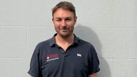 Francis Binney is smiling at the camera. He is wearing a grey polo short with the words marine resources on it in white.