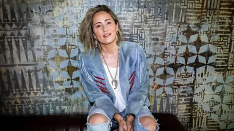 Claire Harrison KT Tunstall in ripped denim jeans and a light blue suede jacket sitting smiling.