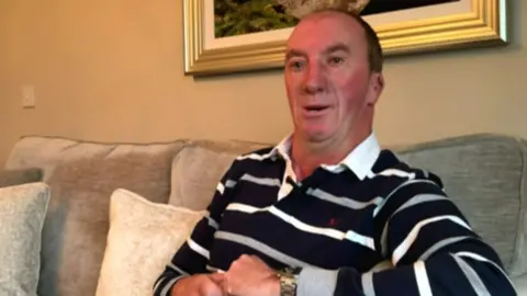 Tom Hadden sits on a grey sofa. He is wearing a stripey top.