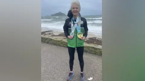 Andrea Simmons stands in front a beach on a choppy day as waves crash onto the sand. The 71-year-old has a Hayle Runners shell suit top on and two large medals around her neck.