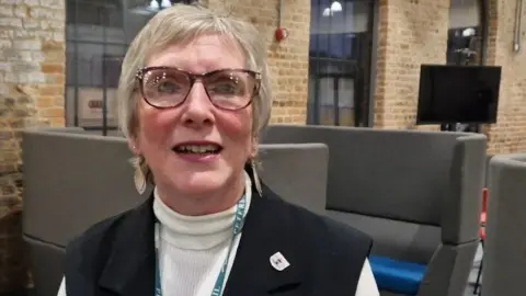 Alison Jones has short fair hair and wears glasses. She is wearing dangly earrings, a cream polo neck jumper and sleeveless black jacket. She is sitting down near banks of soft seating and in a corporate room with exposed bricks and large windows.