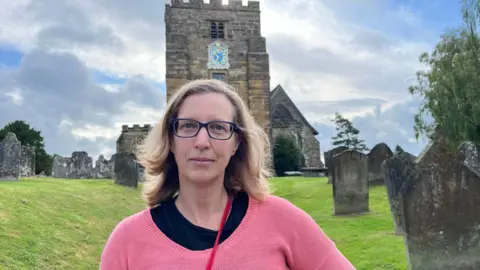 Woman with glasses and pink top stands in front of church in rural village