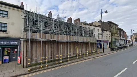 Google A building in a bank of businesses on a main road which has been completely covered with scaffolding and wooden boards.