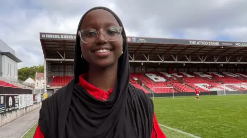 Mariam wears a red City Academy polo shirt, clear glasses and a black hijab. She smiles at the camera from the football pitch, which can be seen behind her.