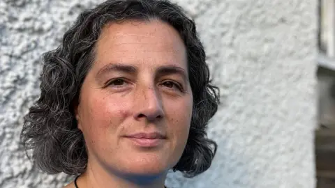 A woman with black and grey curly hair and brown eyes stands in front of a white wall