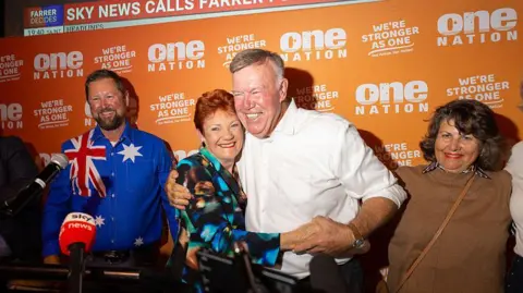 Pauline Hanson wearing a blue shirt hugs David Farley wearing a white shirt with a One Nation sign behind