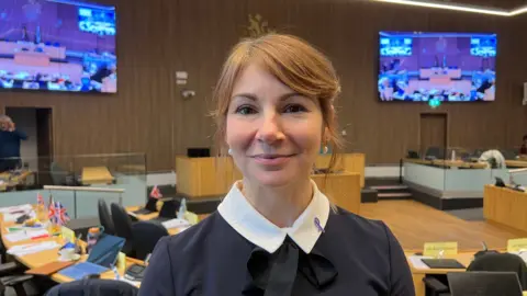 A woman with coppery brown hair smiles softly as she stands in a modern council chambers. Her hair has a sweeping fringe and is tied back. There are big screens with a live feed of the council chambers behind her.