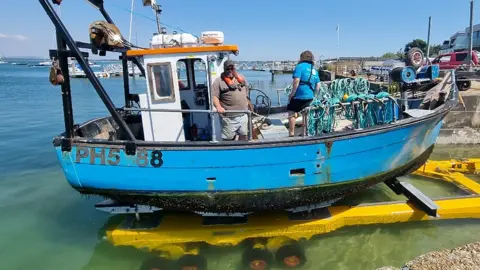 Langstone Harbour Board A small blue fishing boat, with two people on board, sits on a submerged yellow trolley on a slipway