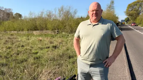 A bald gentleman wearing glasses, a green polo and grey trousers. he is stood on a narrow pavement in between a road and field. 