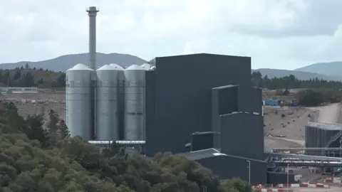 The factory is a large industrial site. There are tall dark grey buildings, several steel silos and chimney-type structure.