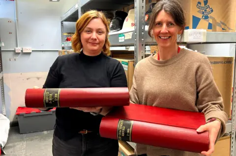 LFB Two smiling women hold large books whilst standing in a storage room.