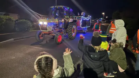 Katie-Marie Fowler A group of children and adults waving at a colourfully decorated grey vintage tractor festooned with fairy lights and pulling a trailer that looks like a sledge containing Santa dressed in traditional red costume with white beard and gloves waving back at the public.