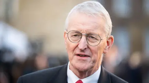 PA Media Hilary Benn has short grey hair and round metal glasses. He is wearing a black suit blazer, white shirt and red tie.