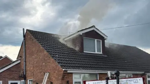 A small amount of smoke rising from a dormer window in the roof of a semi-detached house.