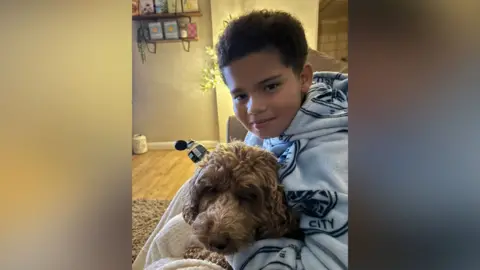 Family A boy with dark brown hair wearing a pale blue Man City blanket hugs his dog on a sofa in a living room. The room has a wooden floor, beige walls and beige rug.