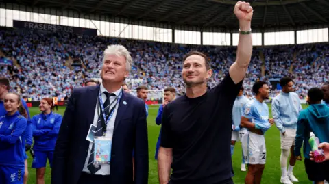 PA Media Two men celebrating on a pitch at a football stadium. The man on the left is wearing a blue suit, white shirt and dark blue tie with grey hair. Next to him is a younger man with brown hair and a blue T-shirt. He is raising his left fist in the air