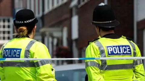 Two police officers in uniform stand with their backs to the camera. One is female the other male. They have blue police tape behind them.