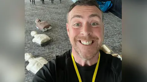 Southport Saviours Foundation Richard Moore with short brown hair and beard wearing a yellow lanyard and black long sleeve top stands with resuscitation manikins on the floor behind him used in a first aid training session. He is smiling. 