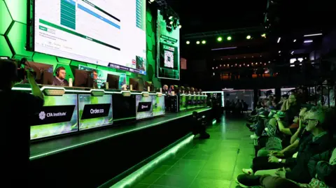 MEWC An arena with a stage on the left and a crowd on the right. The people on the left are sitting at computer desks with large monitors, focusing on Excel challenges with a live stream playing on a giant screen behind them. The crowd on the right, covered in the green light cast by the arena, was watching enthusiastically.