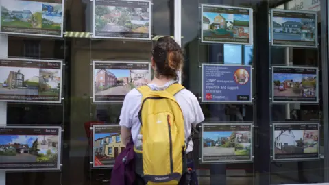 PA Media A woman stands with her back turned looking at the front window of an estate agent.
