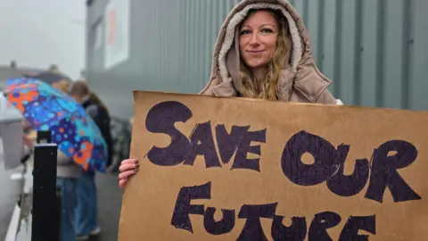 A person stands outside an industrial building in rainy weather holding a cardboard sign that reads “SAVE OUR FUTURE,” with raindrops visible on the surface of the sign.
In the background, other people stand along the roadside with umbrellas and placards, suggesting a small demonstration taking place near the site.