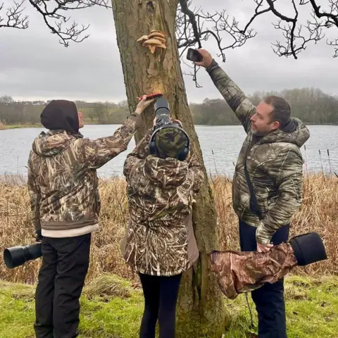Three people wearing camouflage jackets standing outdoors near a loch, examining a tree trunk with several bracket fungi growing on it. Two are holding cameras with large telephoto lenses, and one person is using a smartphone to take a close-up photo of the fungi. The background shows tall dry grass and water under an overcast sky
