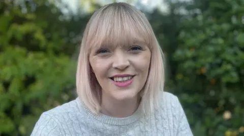 A woman with shoulder length blonde bobbed hair smiles at the camera. She is wearing a grey woollen jumper and is standing in front of greenery