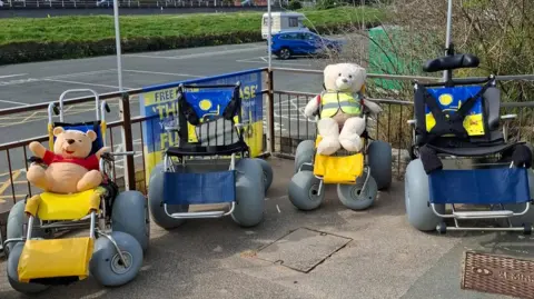 Fleetwood Beach Wheelchairs Four beach buggies with large grey wheels are parked up on a paved area to the front of the organisation's building. Two teddy bears, including Winnie the Pooh, are seated in two of the buggies.