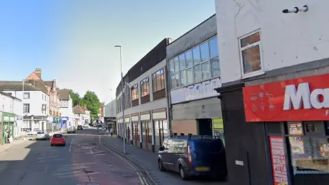 Google There are businesses either side of a road, with the corner of a takeaway with red and white signage visible in the right hand corner of the picture. A dark coloured van is parked on the pavement, and further ahead on the road is a red car and a silver car.