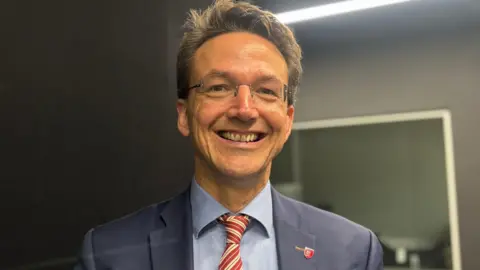 Kirsten Morel has short dark hair and is wearing glasses, a navy jacket, blue shirt, and a stripey red tie.