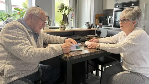 The BBC sits on the table of a marble kitchen in front of an elderly man and a woman in the 60s. Both of them have folders of paperwork and they are looking towards each other. Both are wearing glasses. He has white hair and has brown hair. She is wearing a beige flass and blue trouser, wearing a black and white check trousers and a white arran jumper as well as a large white-teen wrist watch. A window, flower vase, microwave and built-in ovens appear behind them. 