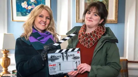 A woman with blonde hair and a woman with brown hair hold a clapper board between them which reads "SHETLAND". Ashley Jenson, the blonde woman, wears a dark green and purple scarf and a dark coat. Alison O'Donnell wears a patterned red scarf, a red jumper and green waterproof jacket. They are inside a formal looking room with baby blue wall panels and white outlines, with pictures hanging on the wall.