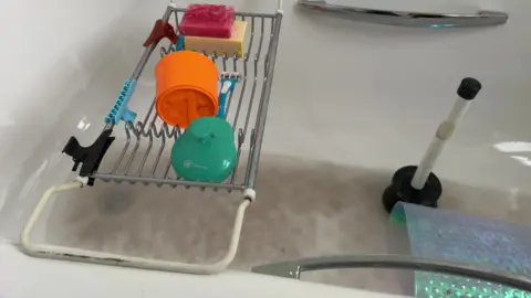 A pool of dirty brown bathwater sits in a white bath in Ms Barley's flat. A black and white plunger can be seen to the right of the photo, and a bath tray sits on top of the bath to the left with hair clips, brushes and soaps on it