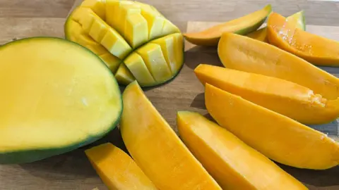 Slices of a Kesar mango on a chopping board next to a standard mango with one side cut into cubes