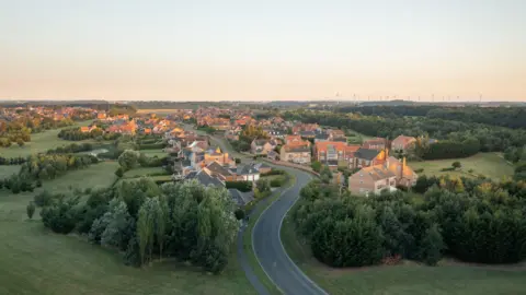 Aerial view of part of Wynyard village. It shows houses on either side of a winding road, with trees on either side. A wind farm can be seen on the horizon.
