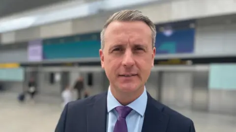 Josie Hannett/BBC Mark Johnston is standing in front of the entrance to Gatwick Airport. He is wearing a dark suit, light blue shirt and purple tie.