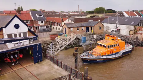 Martin Giles/BBC A bird's eye view of a lifeboat station, with an orange and blue painted boat on the dirty-looking water; rocks on the banks; and industrial and warehouse looking buildings on the land.