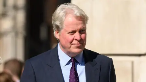 Getty Images Charles Spencer wears a blue suit, blue shirt and purple tie with white spots in it. He is outside, by a large building. He is looking away from the camera.