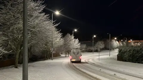 A van on a snow-covered road in Inverness. There is snow lying on the pavements and on trees along the sides of the road. It is dark and the streetlights are on.