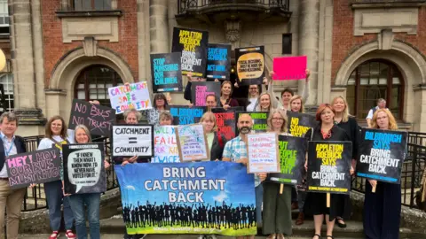 A group of people stood outside an official council building holding up placards. One of the placards read 'Bring catchment back'.