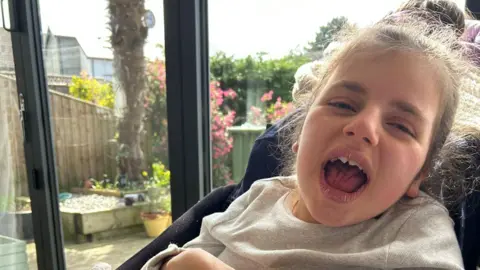 Kirsty Edith sat in a wheelchair by a window looking at the camera with her hair tied back. She is wearing a grey top.