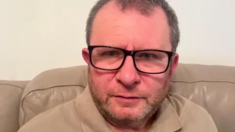 Michael Davies A man in black glasses and a beige t-shirt sitting on a beige leather sofa. A cream wall is behind him.