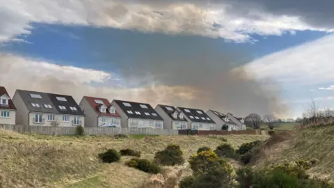 A dark grey plume of smoke rises from the horizon and behind a row of white, harled walled houses.